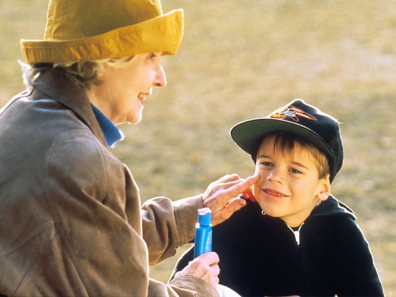 A woman in a yellow hat applying sunscreen to a young boy's face outdoors. The boy is wearing a baseball cap and smiling.