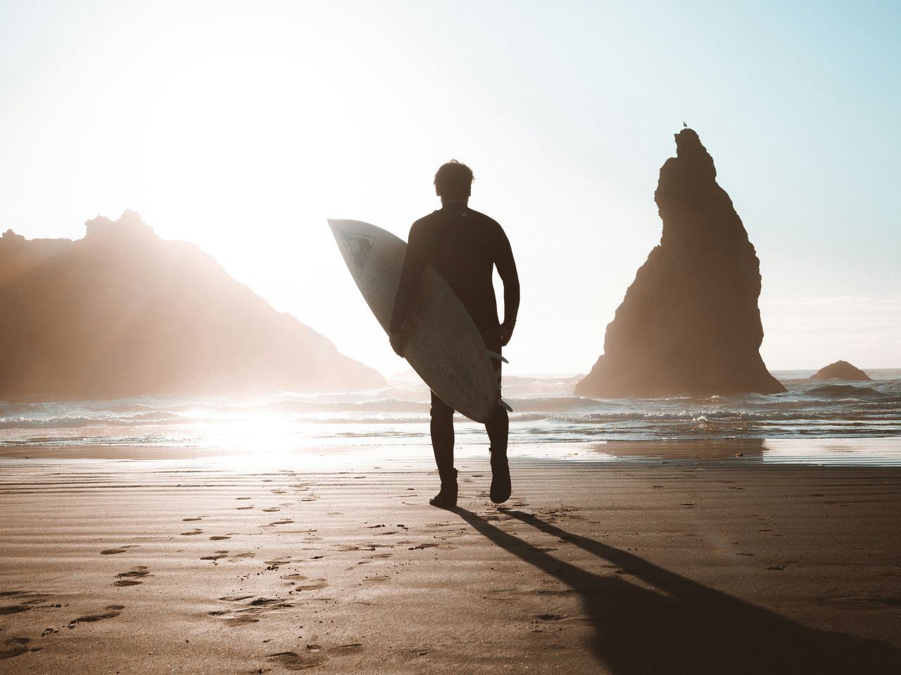 Silhouette of a surfer walking on the beach at sunset or sunrise, carrying a surfboard while approaching the ocean. Dramatic rock formations are visible in the background with golden light creating a beautiful atmospheric scene.